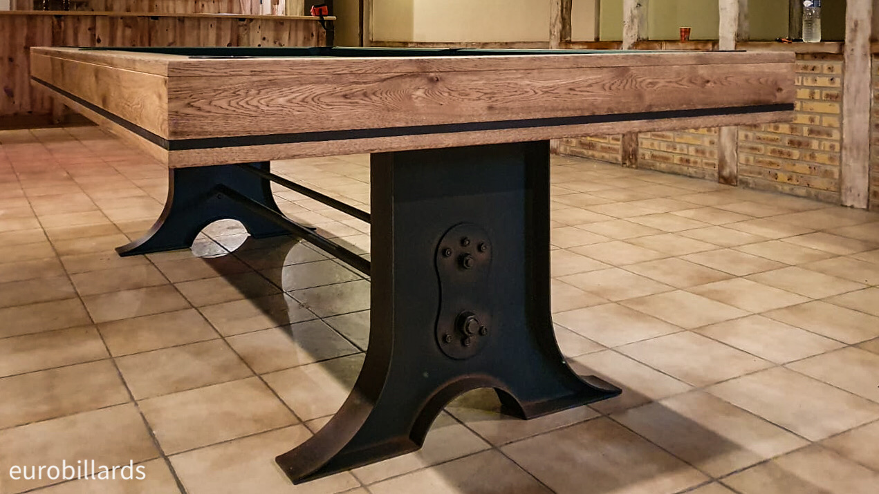 Les gros écrous, les barres métalliques, la forme tulipe et la finition rouille des pieds en métal du billard ATELIER — renforcés ici par le liseré noir sur le cadre en chêne brossé huilé qui soutient le jeu américain au tapis vert épicéa — rappellent les chemins de fer et le style industriel pour prendre naturellement sa place au milieu de cette salle de loisir de la même époque aux murs de briques et poutres apparentes brutes.