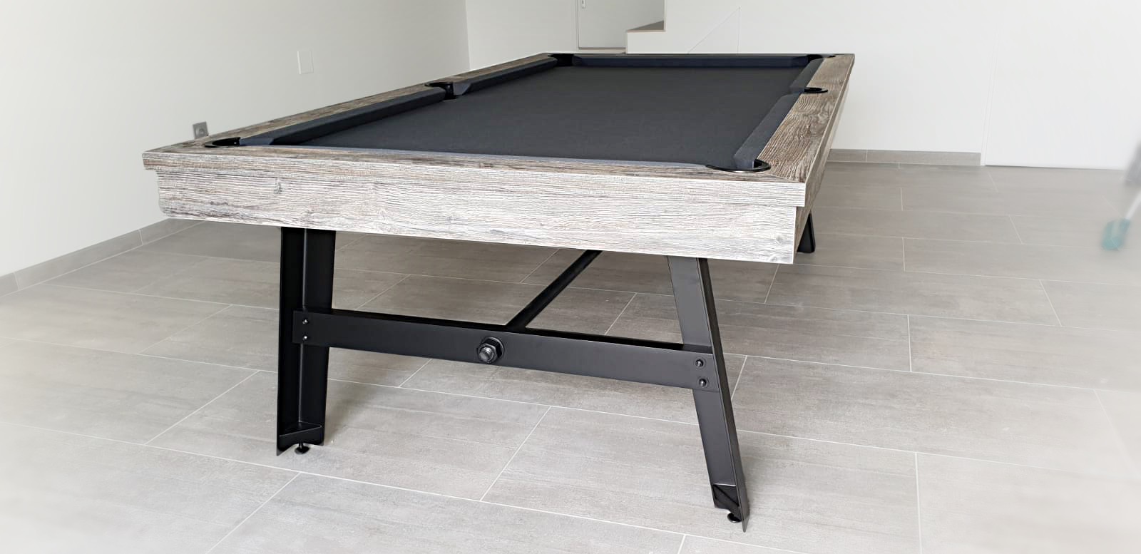 Billard vintage en finition bois flotté sur pieds en acier noir avec son tissu gris anthracite est posé au pied de l'escalier sur le carrelage en grès cérame de cette pièce sobre aux murs blancs immaculés. Parfait dans cette pièce, ce billard ne restera pas au coin.