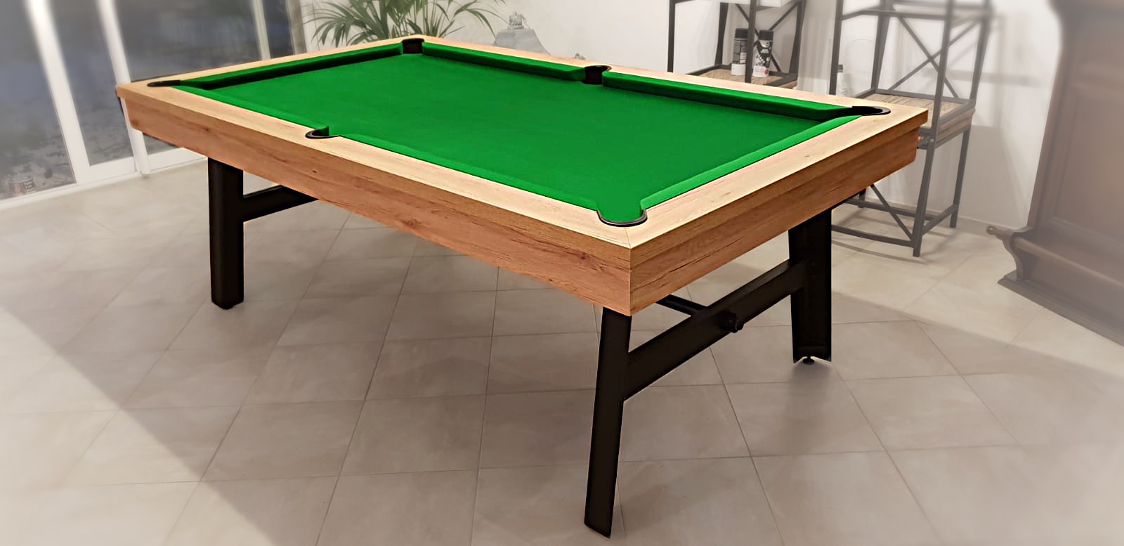 Billard VINTAGE chêne doré veiné sur piétement acier noir avec tissu vert anglais dans une pièce à baie vitrée au carrelage en grès émaillé meublée d'étagères en métal et osier et d'un buffet en noyer avec une plante verte. Son pied industriel au look pliable confère une robustesse à l'image de notre marque de billard.