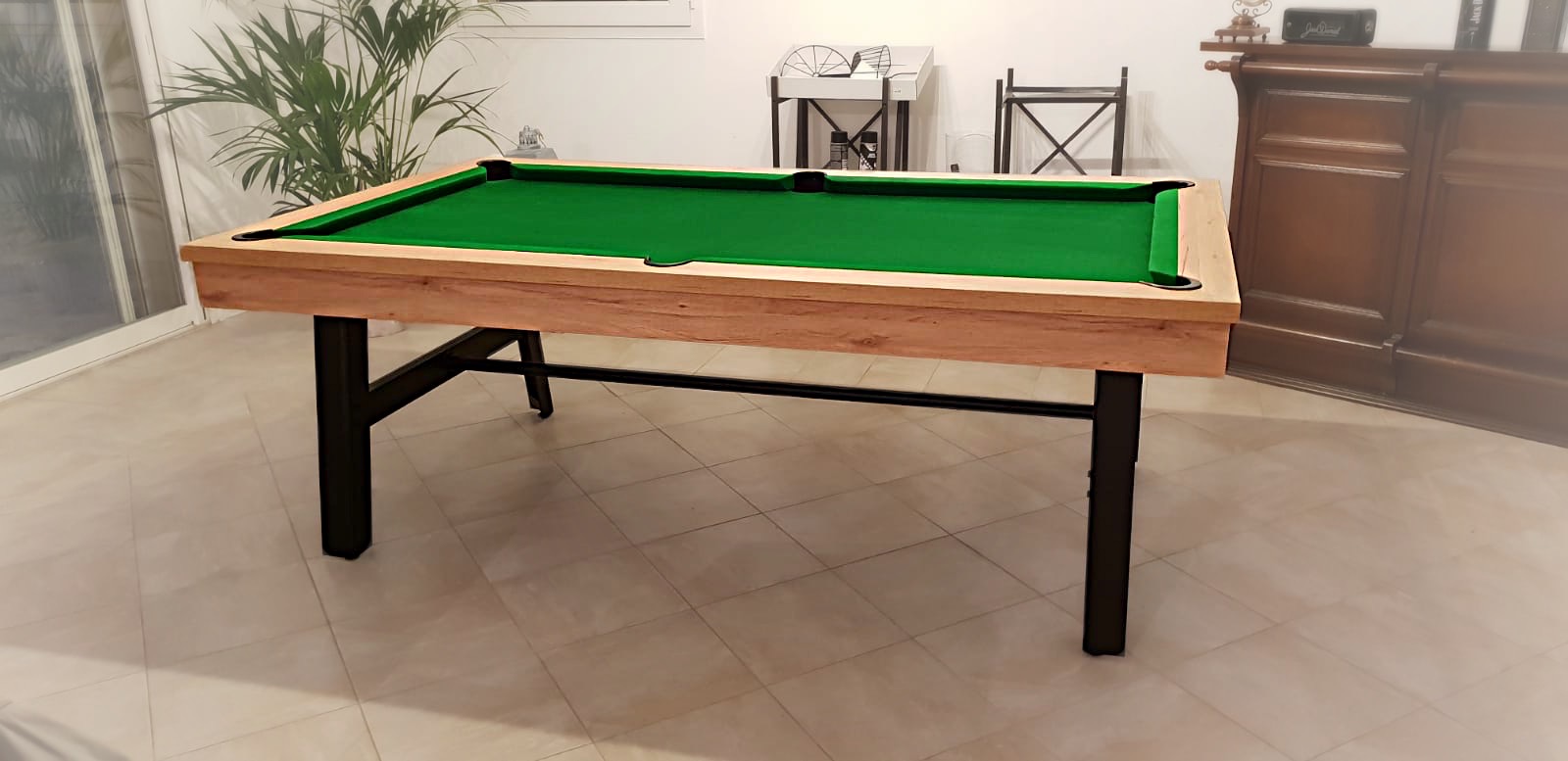 billard vintage en chêne doré veiné de style industriel avec pieds en acier noir et tissu de jeu vert anglais sur carrelage en grès émaillé dans une pièce avec étagères aux pieds en métal fin, buffet en noyer et plante