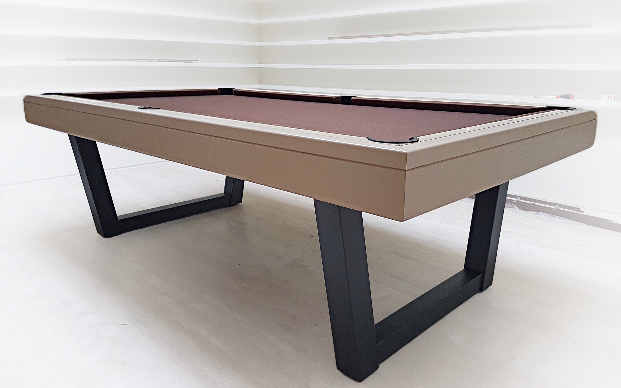 Dans le catalogue de notre marque de billard, ce client a choisi d'acheter un billard HARMONY-V en bois massif laqué RAL 1019 avec un tissu chocolat dont le design en V des pieds gris anthracite en acier inoxydable laqué ressort singulièrement dans sa pièce aux lignes blanches