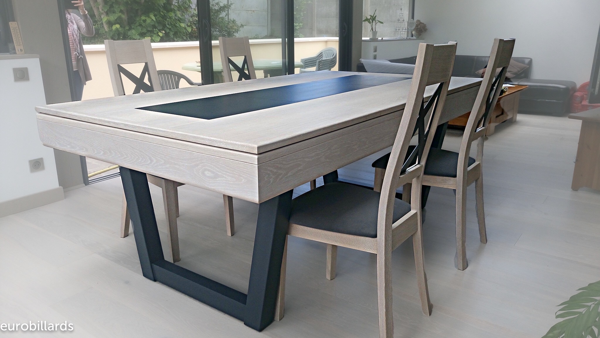 Pour son intérieur au goût moderne, cette famille a choisi le billard HARMONY-V convertible en table dans une finition chêne blanchi avec décor central noir et des pieds en acier inoxydable gris anthracite qui rappellent les montants de leur baie vitrée. Ce modèle phare de la marque de billard française Eurobillards offre un design qui ressort sympathiquement sur le parquet blanc, entouré de chaises Margaux en chêne clair et à l'assise grise également. Superbe optimisation de l'espace à vivre par la double utilisation jeu de billard et de table à manger.