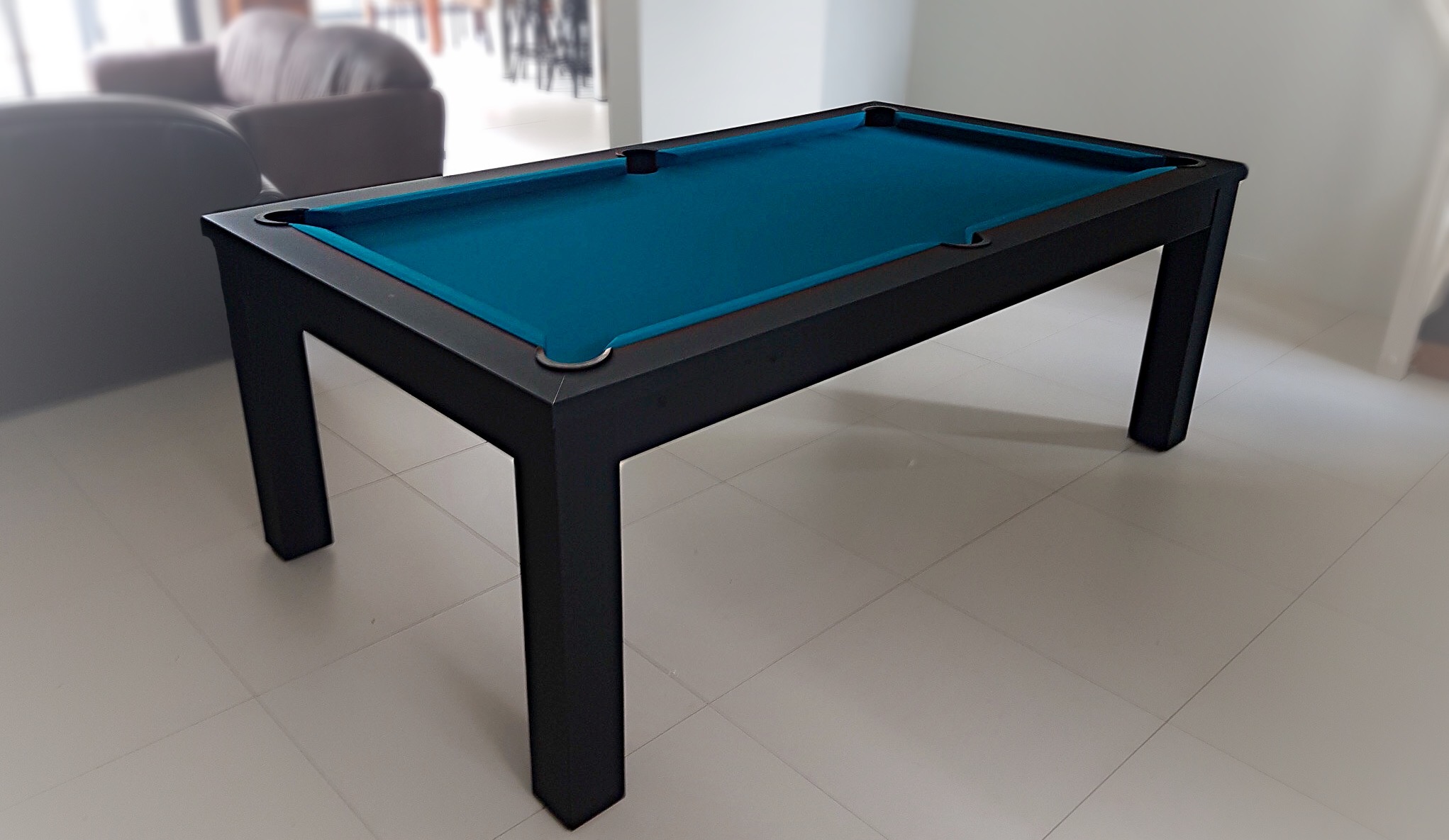 billard trendy noir nano mat en jeu américain au tissu bleu pétrole sur le carrelage émaillé blanc du salon derrière le canapé qui donne sur la baie vitrée de la salle à manger