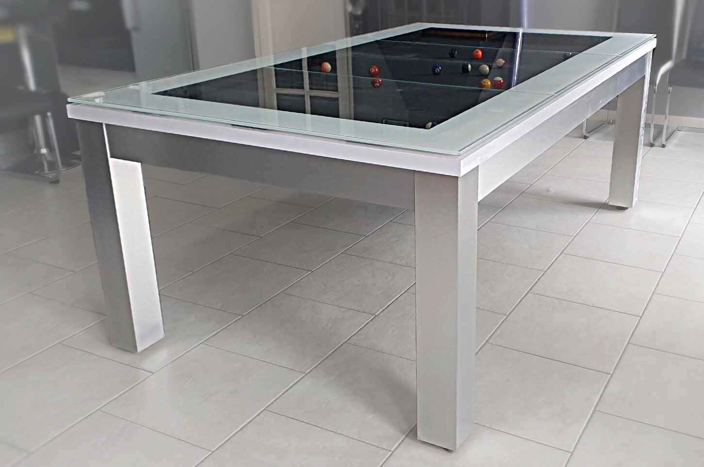 billard trendy au piétement en finition métal brossé sous cadre blanc brillant surmonté d'un plateau VR aui montre le jeu américain au tissu gris ardoise qui fait écho aux chaises à l'assise grise et au piétement métallique dans cette pièce de vie au carrelage émaillé blanc et aux portes vitrées