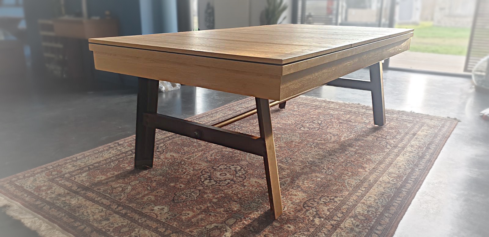 billard transformable en table vintage chêne nature sur pied en acier vieilli rouillé avec plateau ST sous un rayon de lumière et sur un tapis oriental qui recouvre un sol béton ciré billard transformable en table vintage chêne nature sur pied en acier vieilli rouillé avec plateau ST sous un rayon de lumière et sur un tapis oriental qui recouvre un sol béton ciré